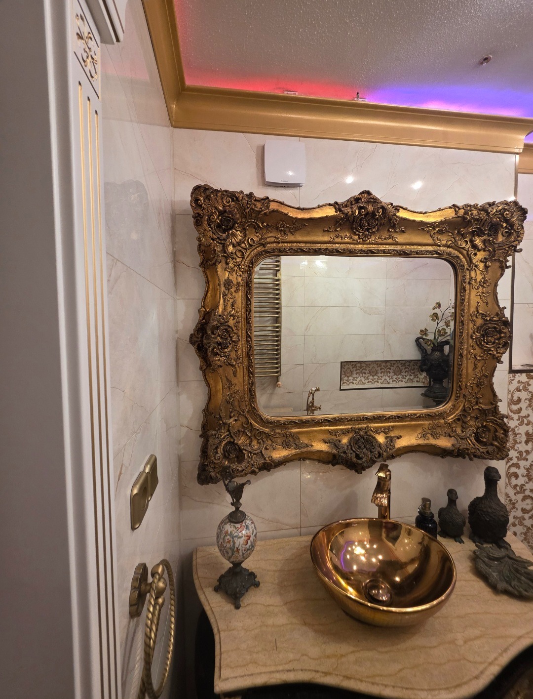 Złota umywalka i bogato zdobione lustro w łazience w stylu glamour. Marmurowe ściany i blaty, dekoracyjne akcesoria. Oświetlenie sufitowe podkreśla luksusowy charakter.