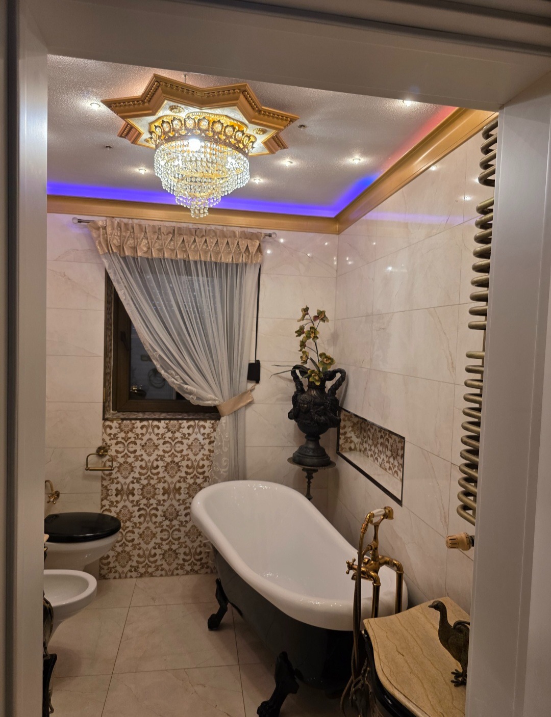 Luksusowa łazienka w stylu glamour: wolnostojąca wanna na lwich nóżkach, kryształowy żyrandol i marmurowe płytki. Dekoracyjny grzejnik drabinkowy i złote akcenty.