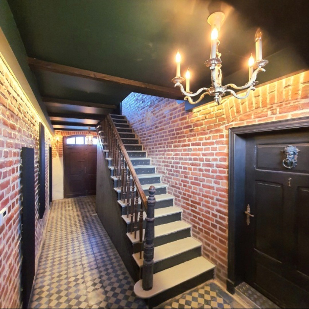 Korytarz z ceglanymi ścianami, schodami z drewnianą balustradą i szachownicową podłogą. W tle widoczne drzwi wejściowe. Całość oświetla ozdobny żyrandol.