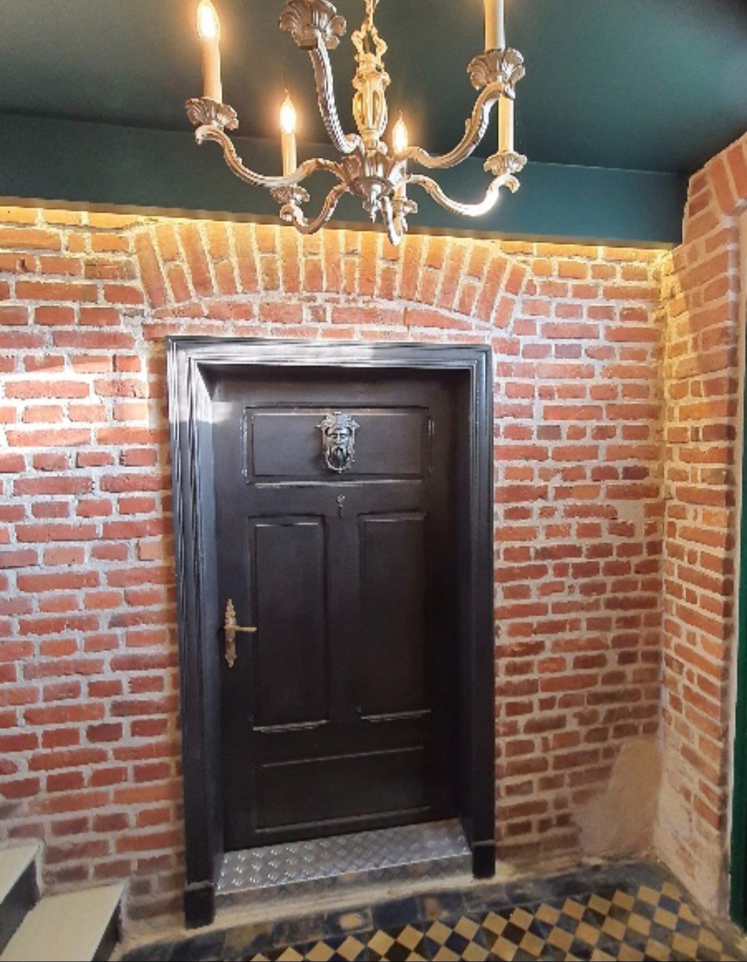 Ciemne drzwi z kołatką w kształcie lwa, wkomponowane w ścianę z czerwonej cegły. Nad drzwiami łuk z cegieł i ozdobny żyrandol.