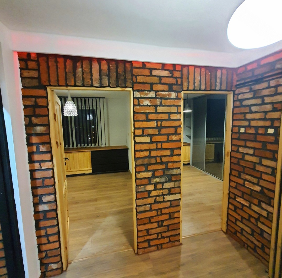 Wykończenie wnętrza: ściany i obramowania drzwi z cegły klinkierowej, drewniana podłoga. Widok na pomieszczenia z meblami i oknem z żaluzjami w tle.