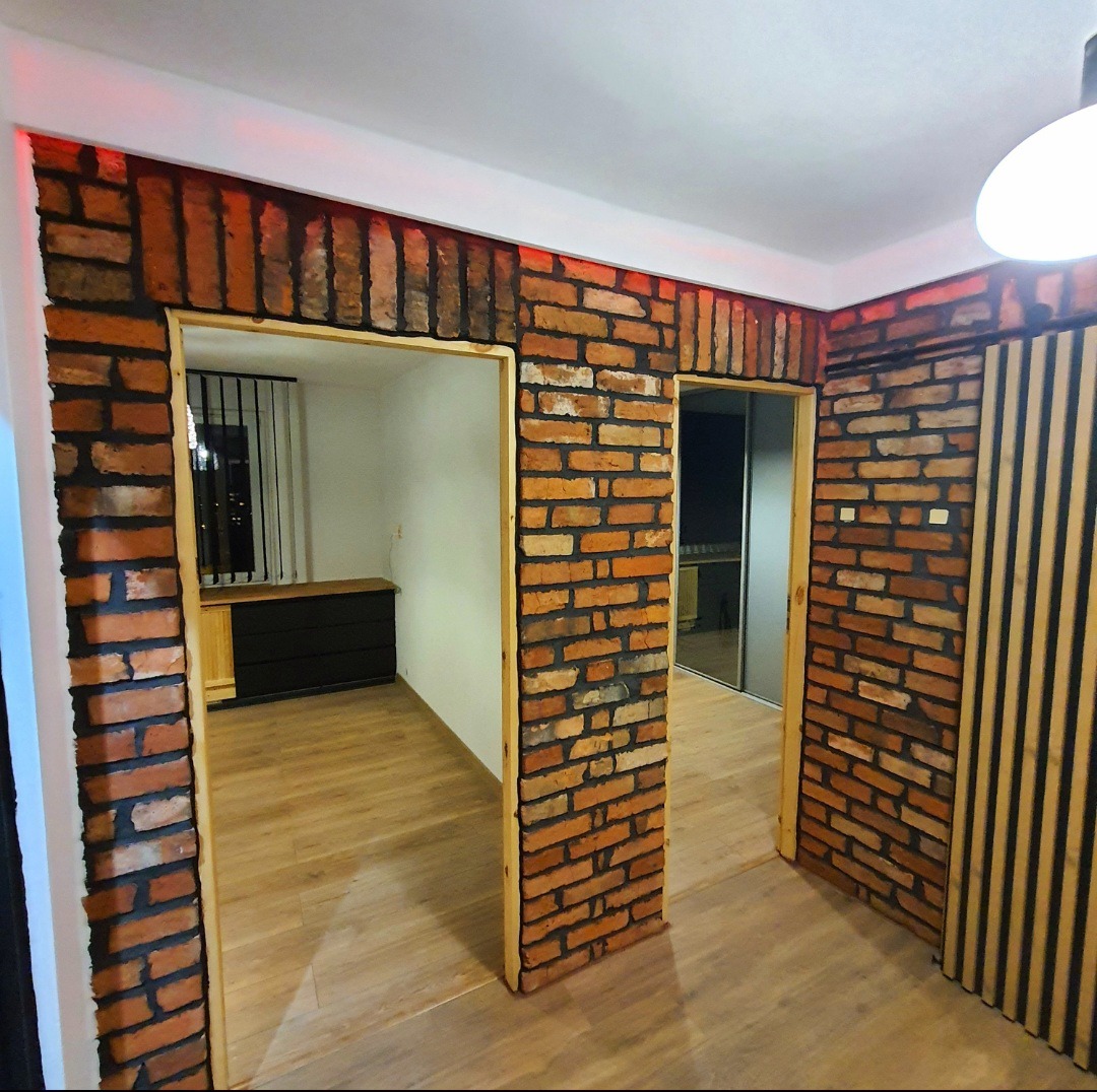 Wykończenie wnętrza: ściany z cegły klinkierowej z drewnianymi listwami, oświetlenie LED, widok na parkiet i meble przez dwa otwory drzwiowe.