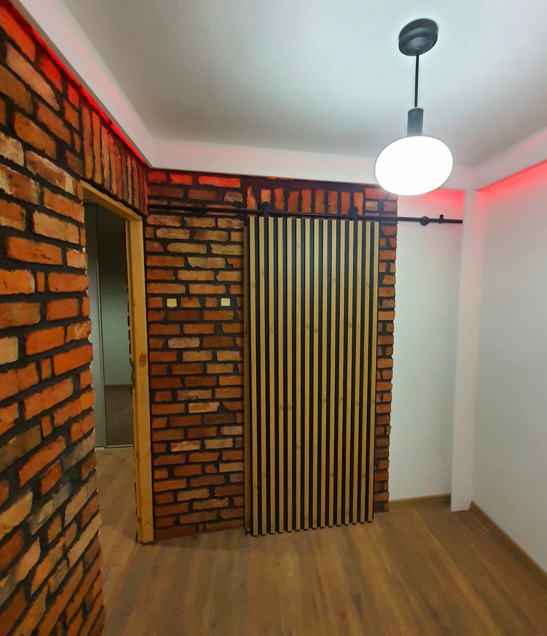 Loftowe wnętrze: ściana z cegły, drewniana okładzina lamelowa, parkiet i designerska lampa sufitowa. Czerwone oświetlenie LED podkreśla surowy charakter.