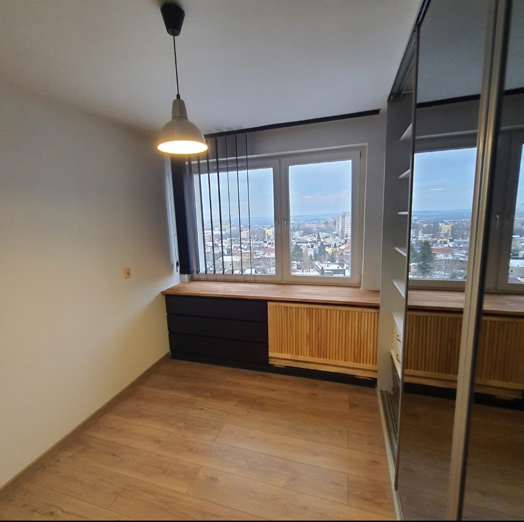 Jasne wnętrze pokoju z drewnianą podłogą, zabudową okienną z grzejnikiem i szafą z lustrzanymi drzwiami. Widok na miasto z okna.