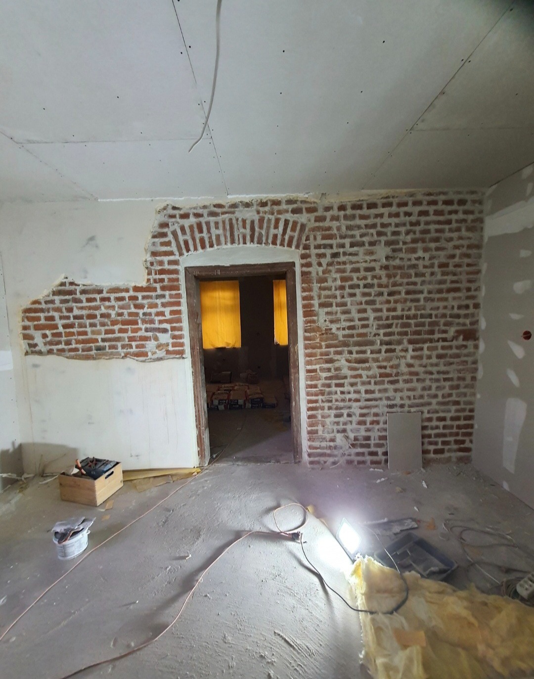 Pokój w trakcie remontu: odsłonięta cegła na ścianie z łukowym przejściem, surowy beton na podłodze, oświetlenie budowlane. Widoczne narzędzia i materiały.
