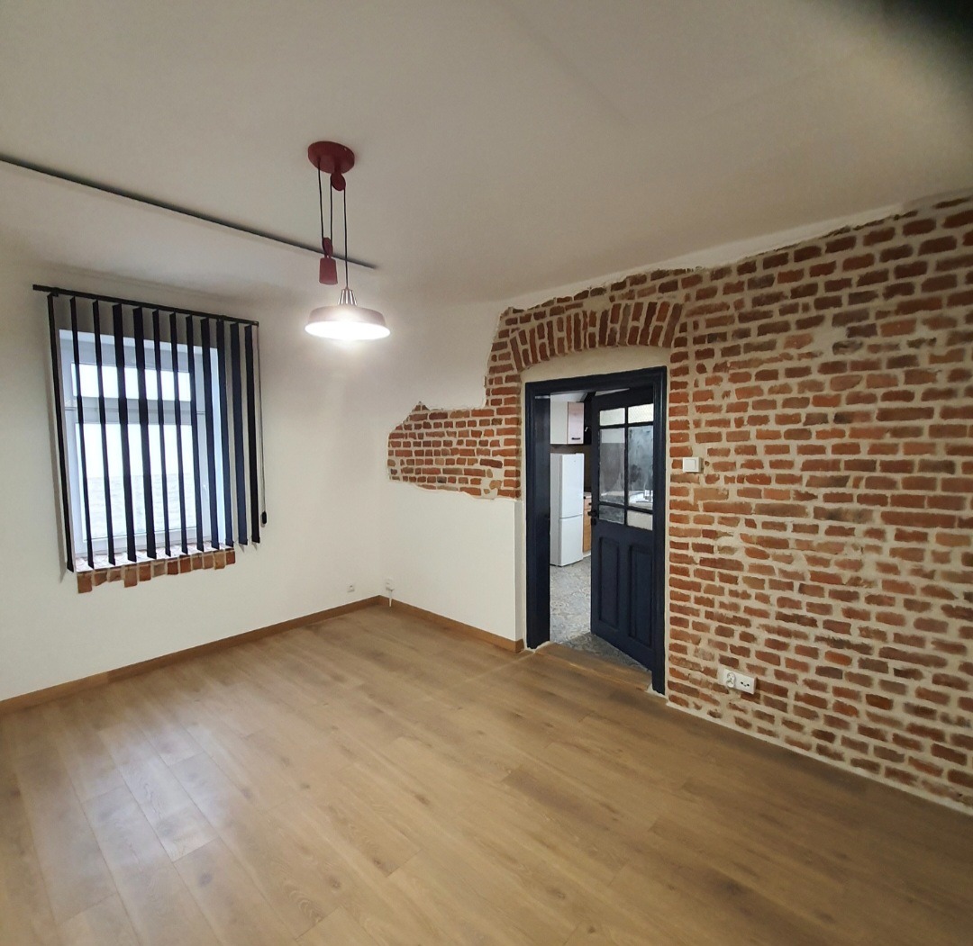Wnętrze po renowacji: parkiet, ceglana ściana z ościeżnicą, okno z żaluzjami pionowymi, lampa wisząca. Widok na kuchnię. Charakterystyczne wykończenie ściany.