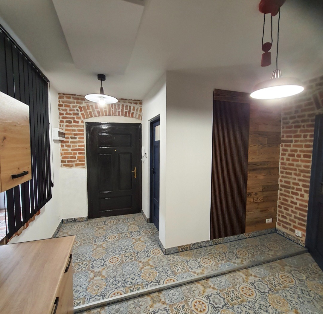 Wnętrze po remoncie: wzorzysta podłoga, ceglane ściany, drewniane panele i czarne drzwi. Nowoczesne oświetlenie. Widok na korytarz i wejście.