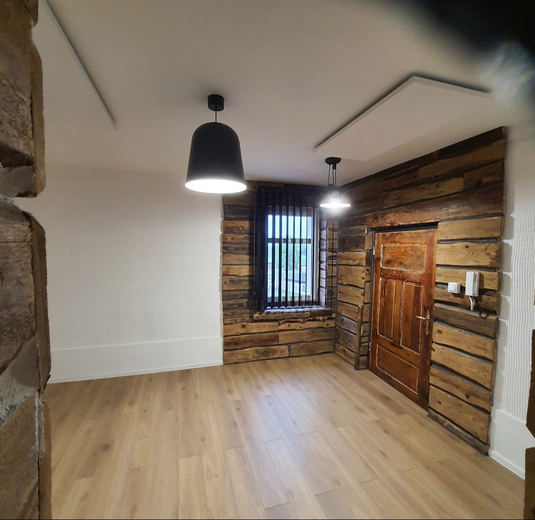 Jasne wnętrze z drewnianymi ścianami i panelami podłogowymi. Okno z żaluzjami i dwoje drzwi. Dwie lampy sufitowe oświetlają przestrzeń. Ściana pomalowana na biało.