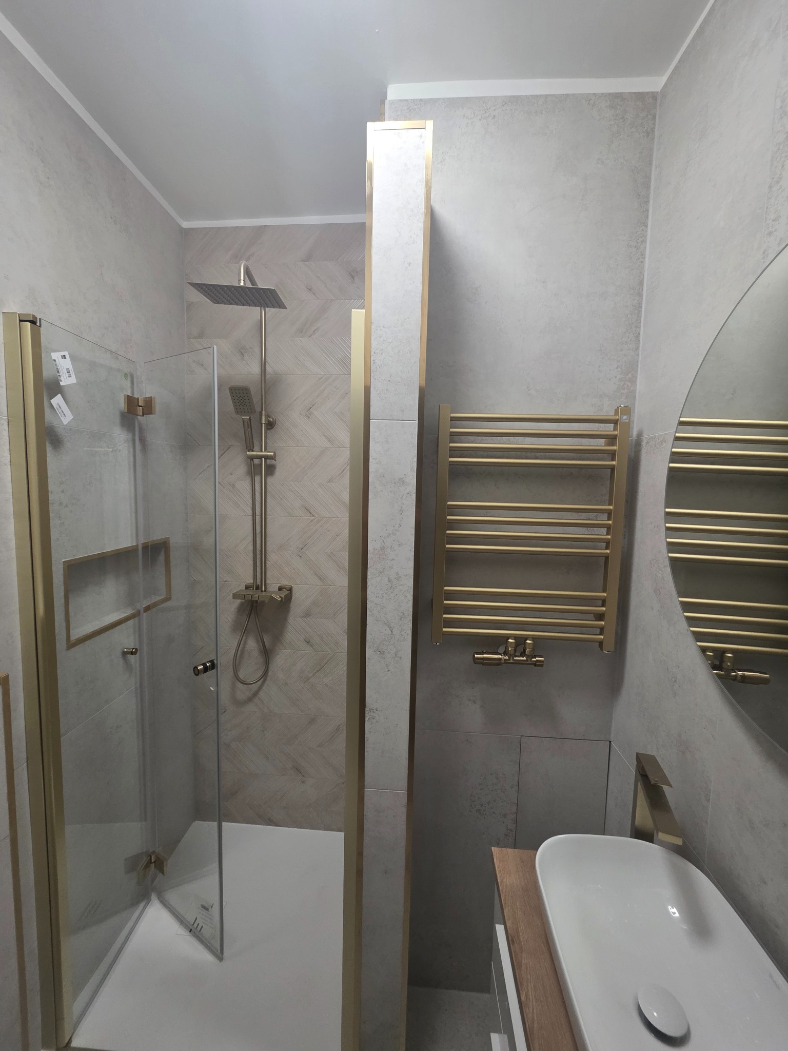 Nowoczesna łazienka z kabiną prysznicową typu walk-in, grzejnikiem drabinkowym i umywalką nablatową. Armatura w kolorze złotym, płytki w odcieniach szarości.