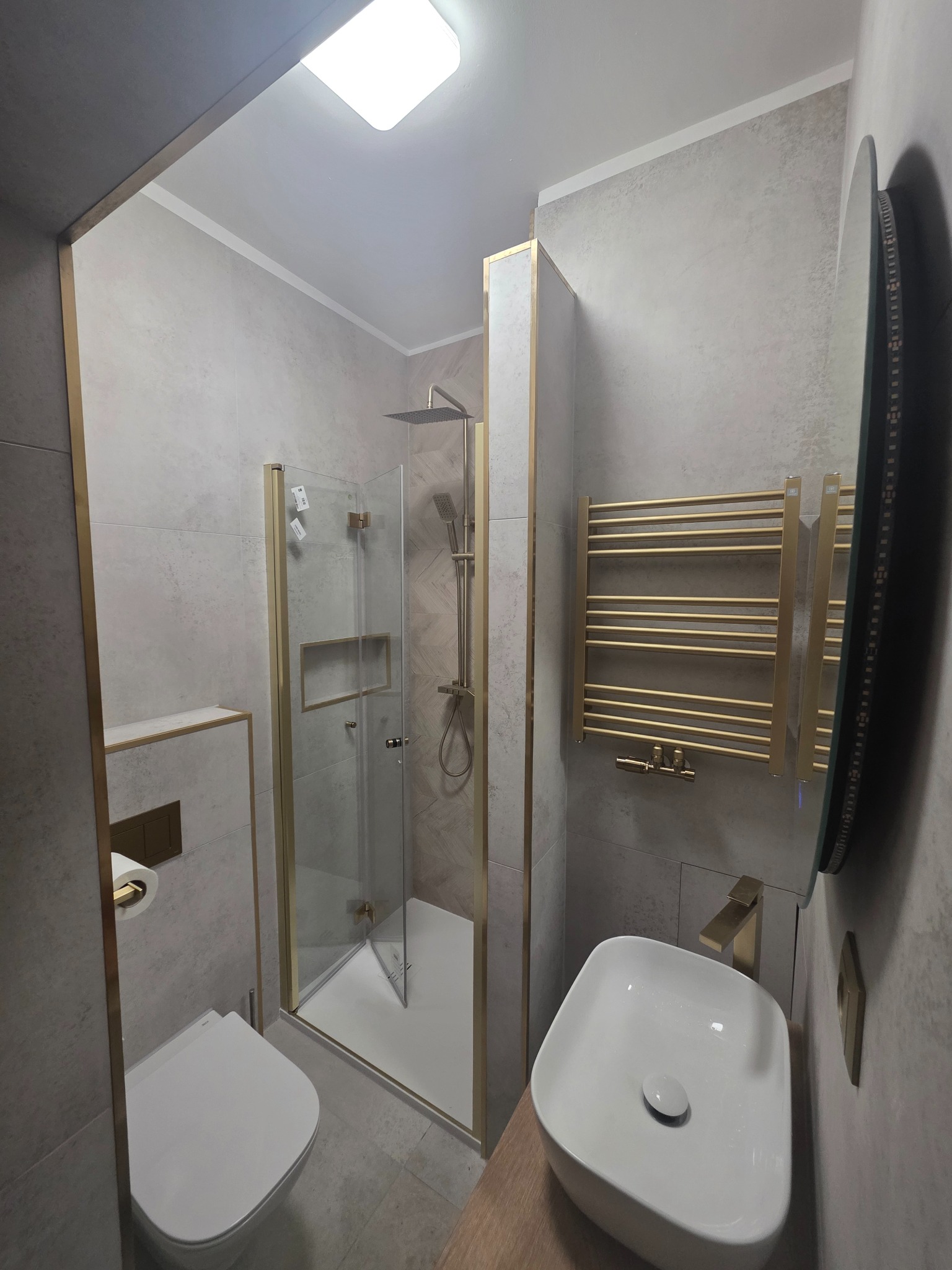 Nowoczesna łazienka po remoncie: kabina prysznicowa ze złotymi ramami, umywalka nablatowa, grzejnik drabinkowy w kolorze złota i podwieszana toaleta.