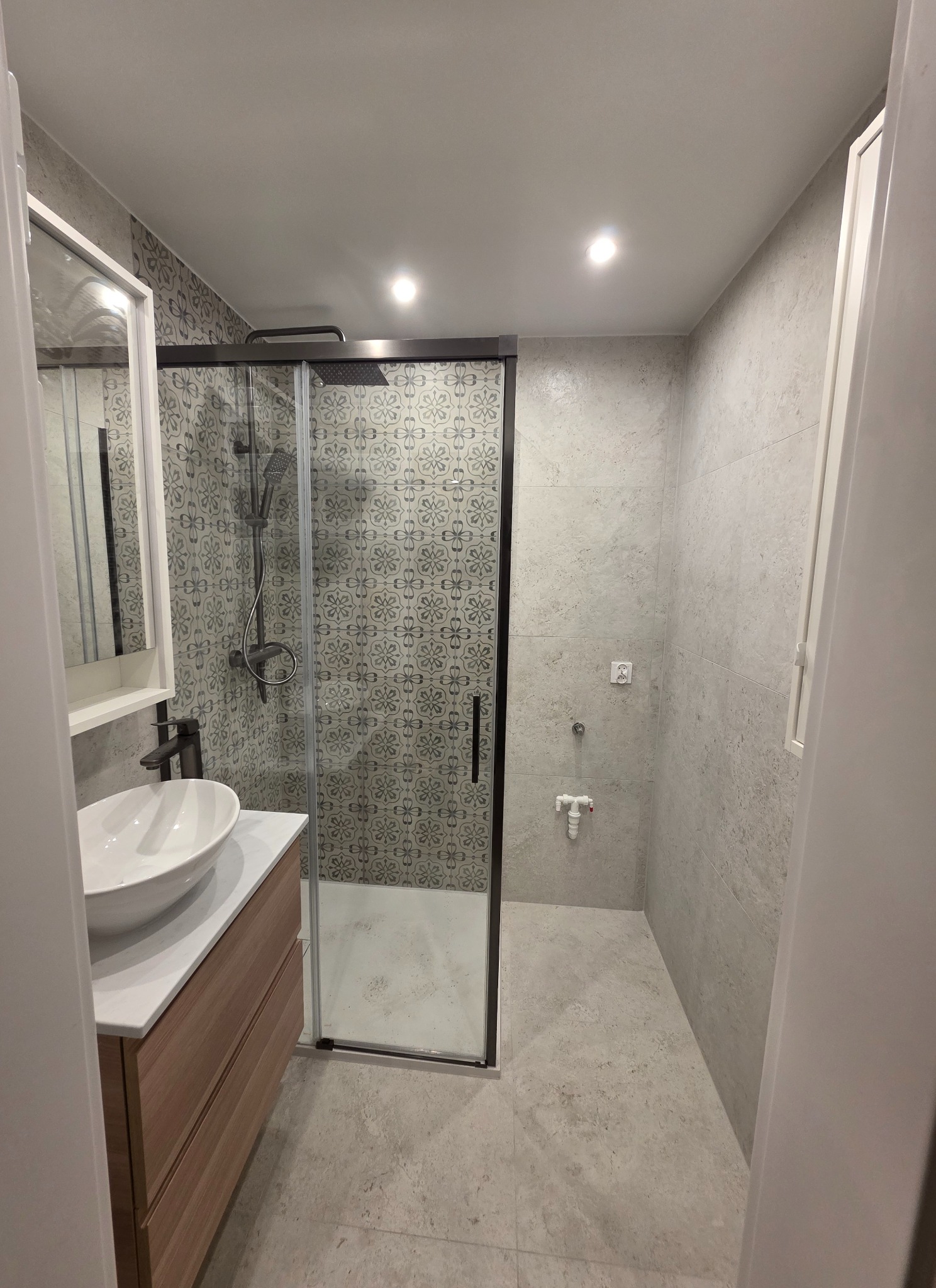 Nowoczesna łazienka z kabiną prysznicową typu walk-in, umywalką nablatową na drewnianej szafce i szaro-białymi płytkami z wzorem. Minimalistyczny design, jasne oświetlenie.