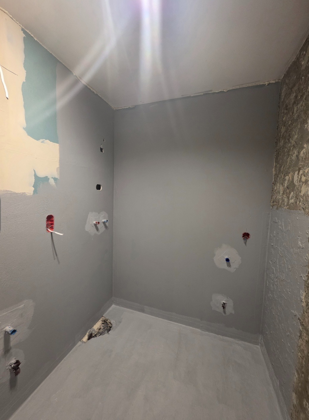 Szara łazienka w trakcie remontu. Widoczne przygotowane ściany, zagruntowana podłoga i wystające rury. Fragment ściany z odsłoniętym murem. Otwory na instalacje.