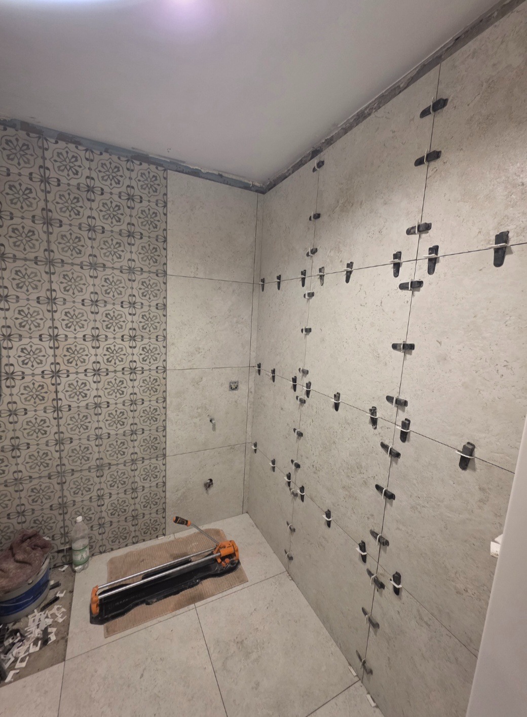 Łazienka w trakcie prac: częściowo ułożone płytki z wzorem, reszta ścian w jasnych płytkach z systemem poziomowania. Na podłodze gilotyna do cięcia płytek.