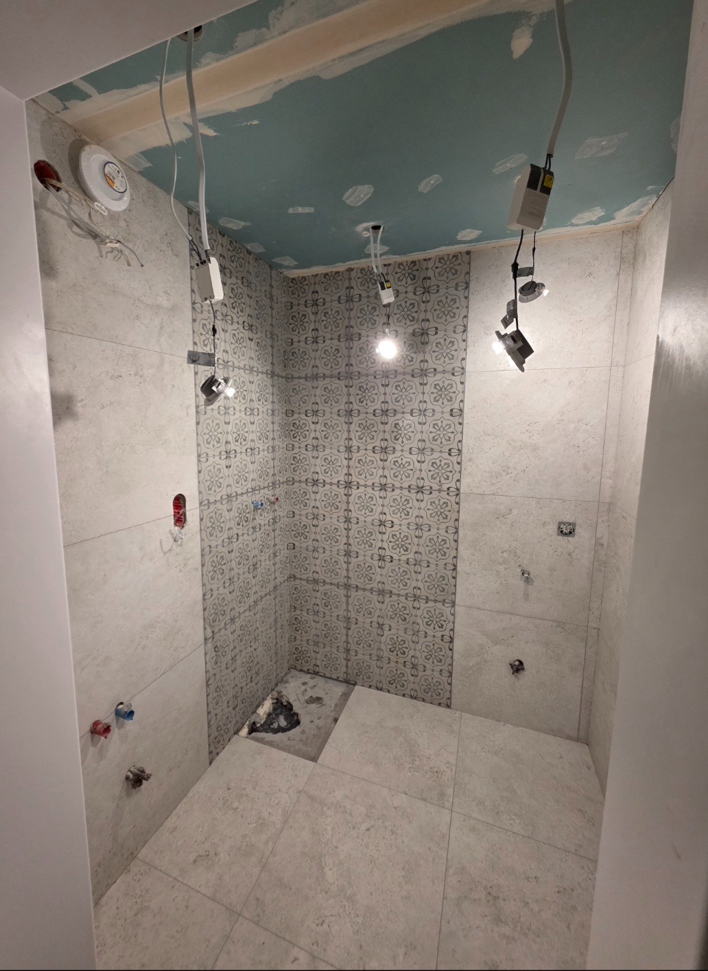 Wnętrze łazienki w trakcie wykańczania: ułożone płytki ścienne z wzorem, częściowo ułożona podłoga, widoczne instalacje elektryczne i hydrauliczne, sufit w trakcie malowania.