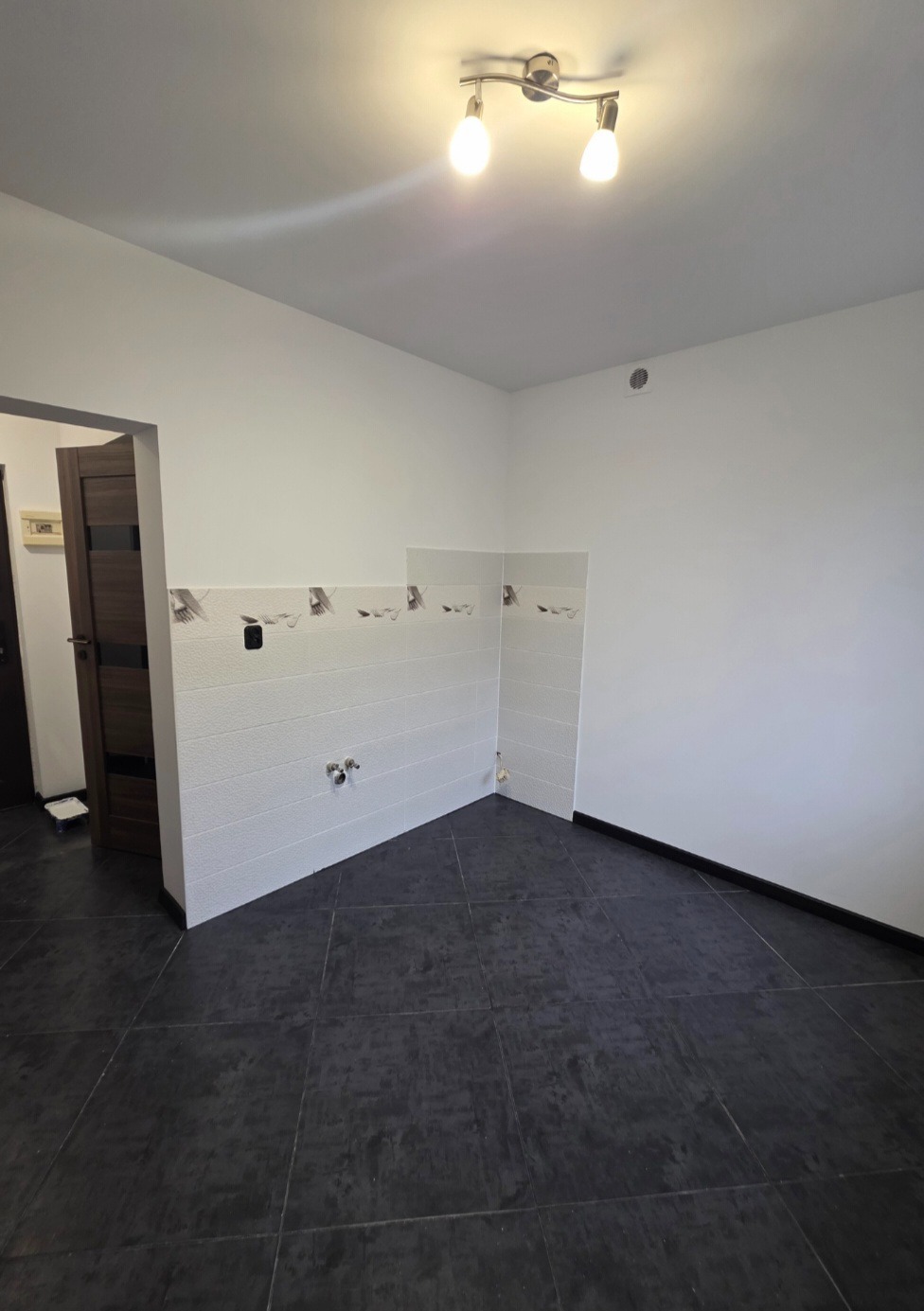 Wykończona wnęka kuchenna z białymi płytkami z dekoracyjnym paskiem i czarną podłogą. Widoczne przyłącza wodne. Oświetlenie sufitowe.