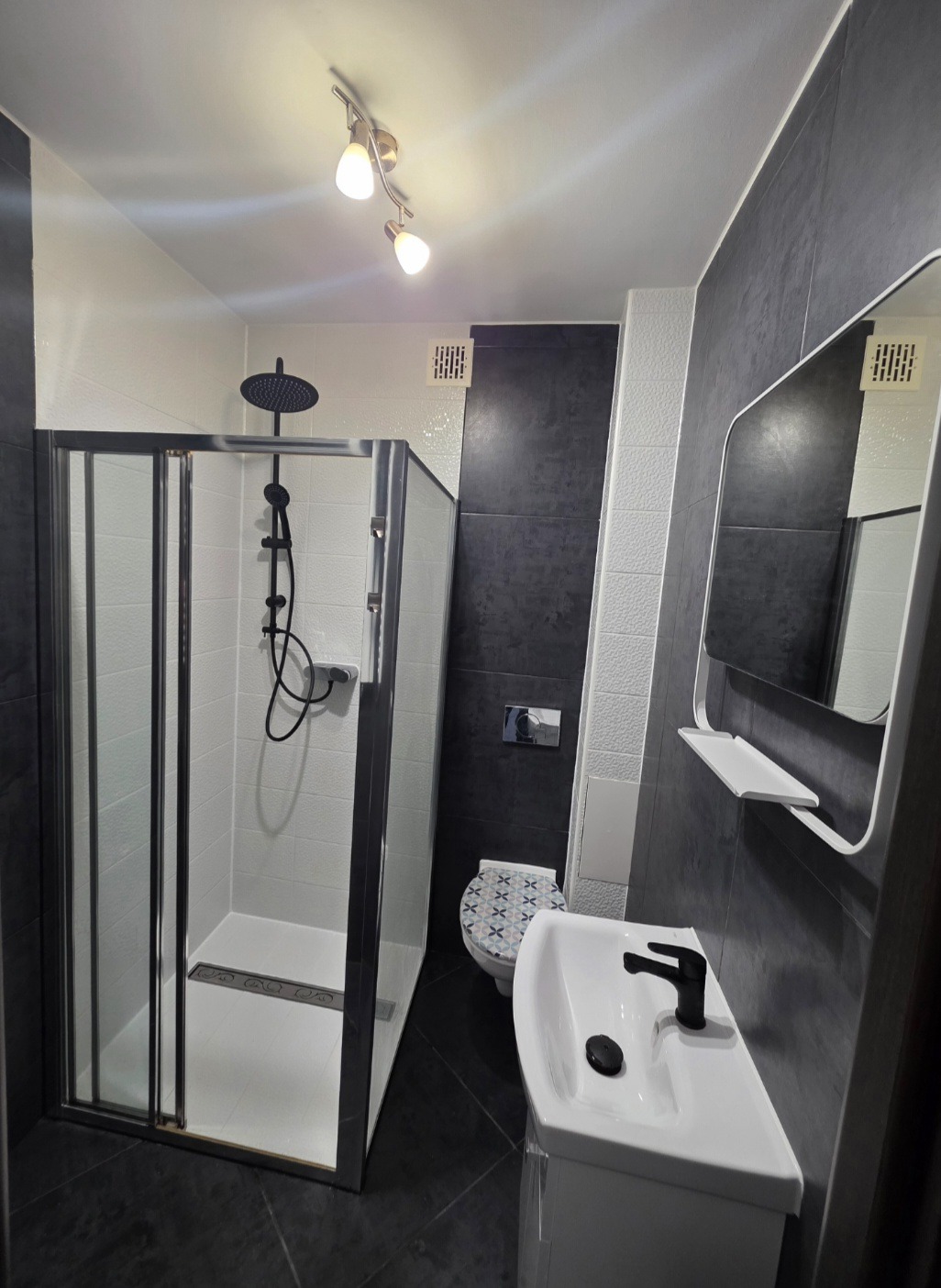 Nowoczesna łazienka po remoncie: kabina prysznicowa z czarną armaturą, umywalka z szafką, lustro z półką i toaleta w odcieniach szarości i bieli. Oświetlenie sufitowe.