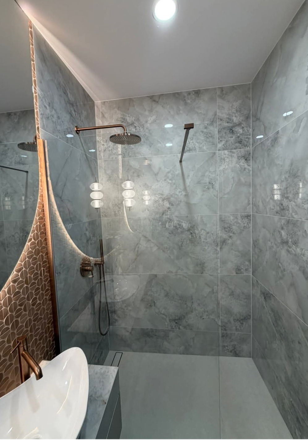 Nowoczesna kabina prysznicowa z szarymi płytkami imitującymi marmur, deszczownicą w kolorze miedzi i dekoracyjnym lustrem z podświetleniem LED.