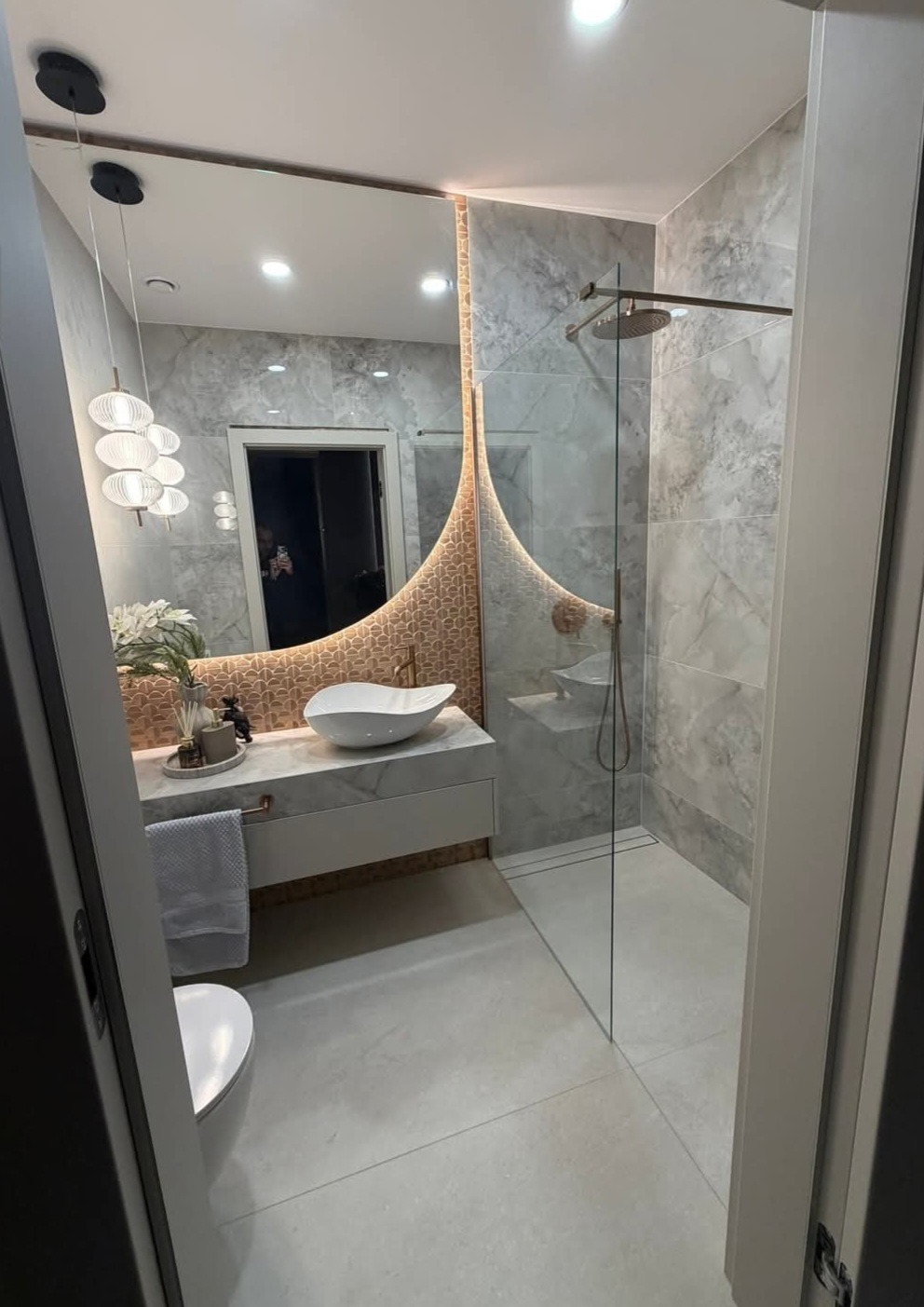 Nowoczesna łazienka z marmurowymi płytkami, owalnym lustrem z podświetleniem i szklaną kabiną prysznicową typu walk-in. Uwagę przykuwa designerska umywalka nablatowa.