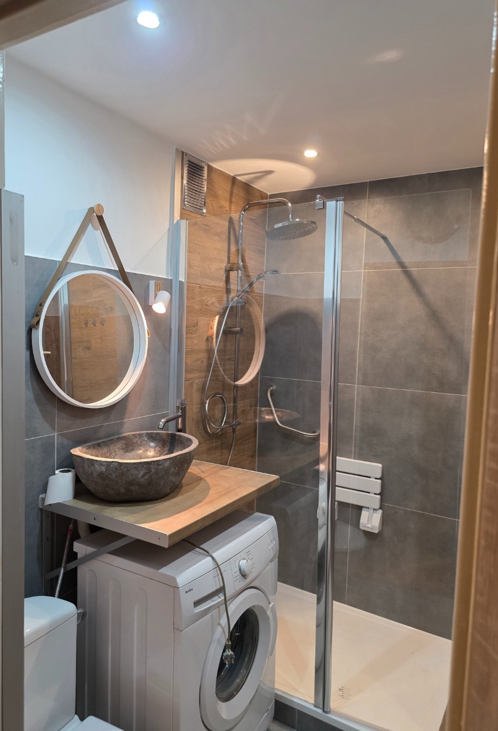 Nowoczesna łazienka z kabiną prysznicową, umywalką w stylu kamiennej misy i okrągłym lustrem na tle szarych płytek. Pralka pod blatem umywalki.