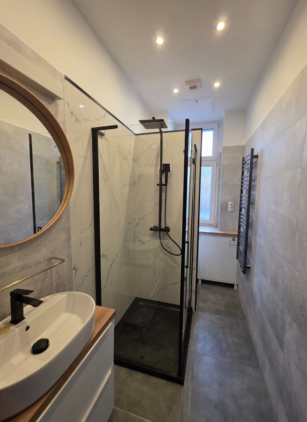 Nowoczesna łazienka z kabiną typu walk-in z czarnymi profilami, okrągłym lustrem w drewnianej ramie i umywalką nablatową. Szare płytki na ścianach i podłodze.