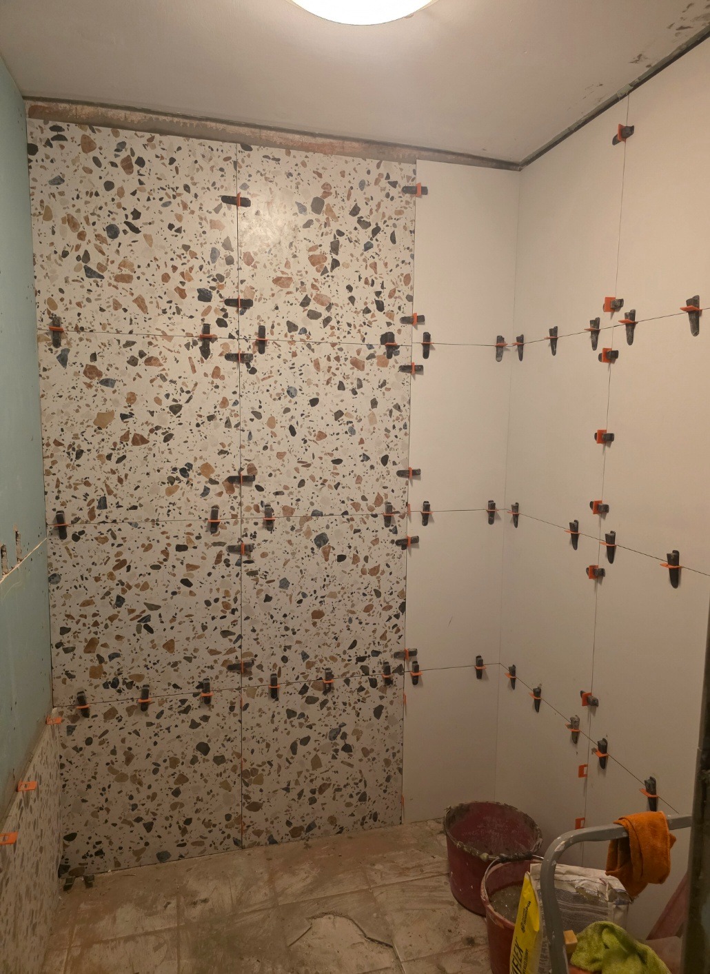 Ściana łazienki w trakcie układania płytek imitujących lastryko, z użyciem klipsów poziomujących. Część ściany wykończona gładkimi, białymi płytkami.