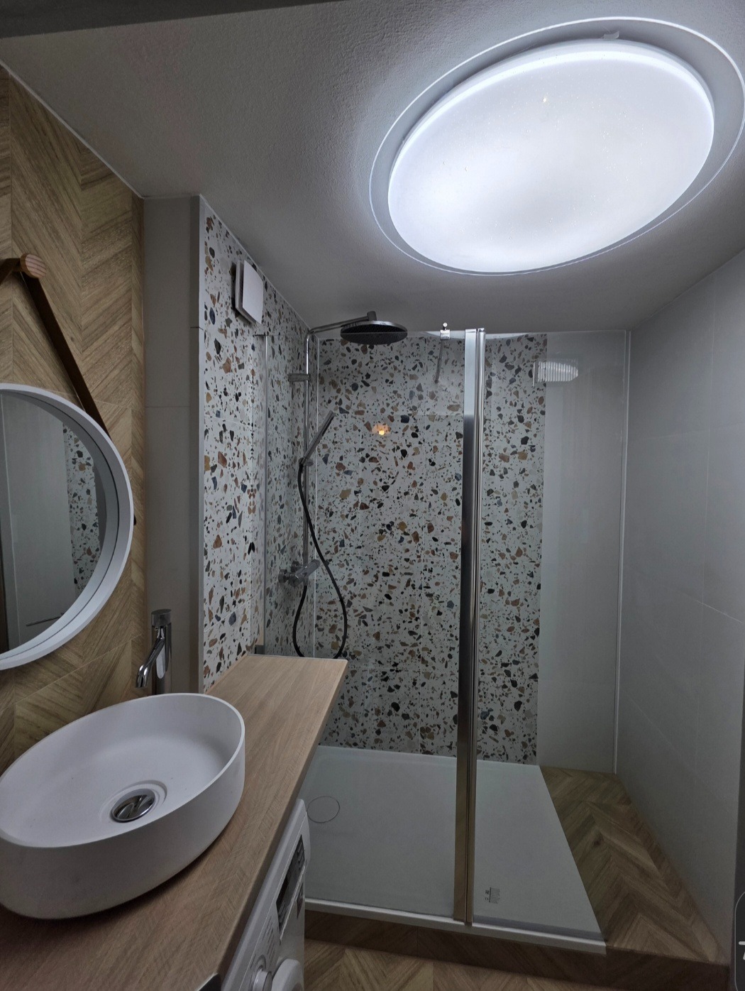 Nowoczesna łazienka z kabiną prysznicową typu walk-in, umywalką nablatową i oryginalną mozaiką na ścianach. Oświetlenie sufitowe w kształcie okrągłego panelu LED.