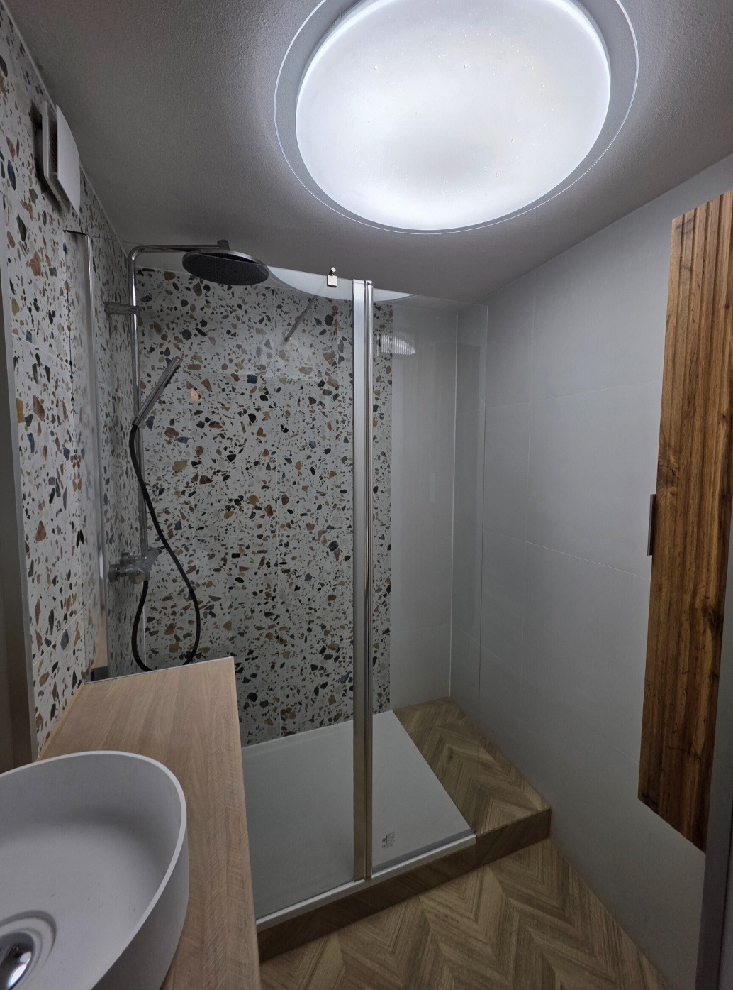 Nowoczesna łazienka z kabiną prysznicową typu walk-in, wykończona płytkami lastryko i drewnianymi akcentami. Widoczne oświetlenie sufitowe i umywalka.