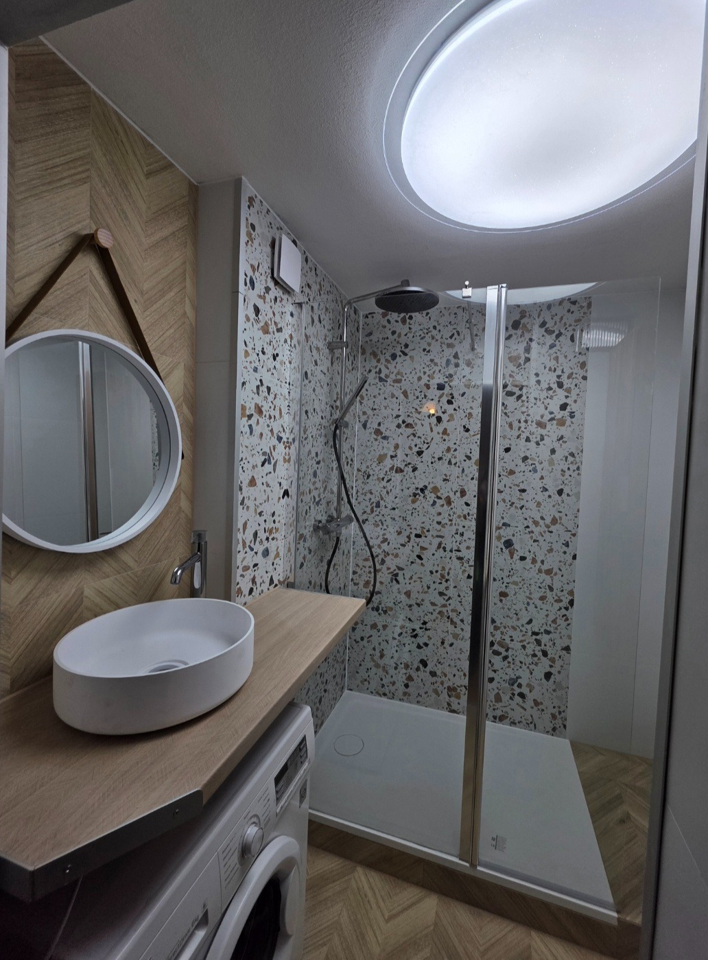 Nowoczesna łazienka z kabiną prysznicową i umywalką nablatową na drewnianym blacie, pod którym znajduje się pralka. Ściany wyłożone płytkami w mozaikę.