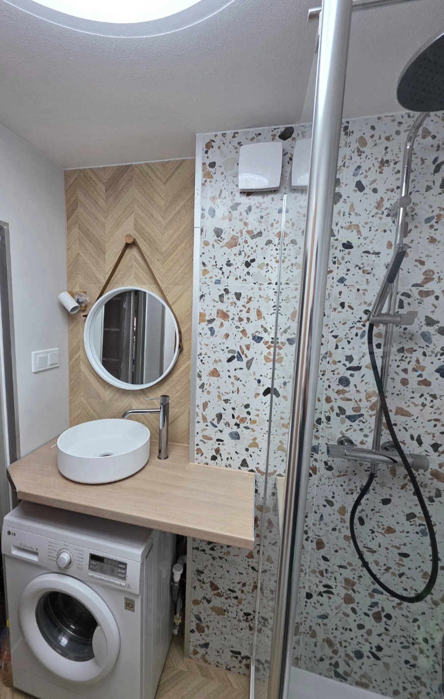 Nowoczesna łazienka z umywalką nablatową na drewnianym blacie nad pralką, okrągłe lustro na ścianie w jodełkę i kabina prysznicowa z mozaiką.