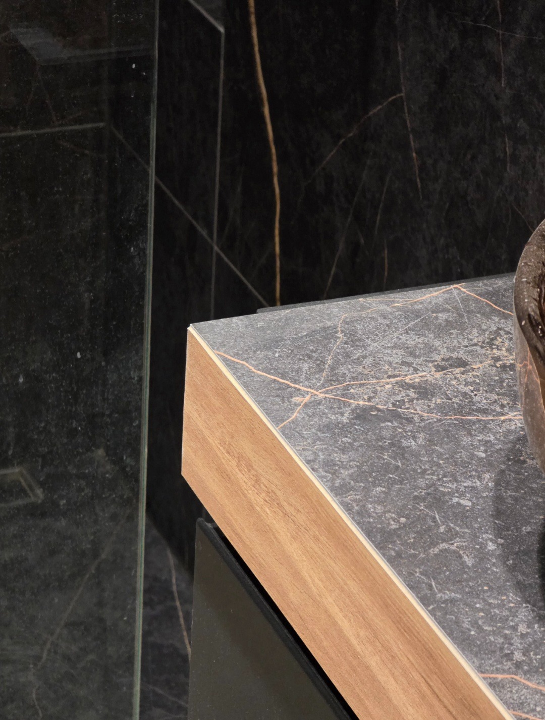 Detal wykończenia łazienki: blat z szarego marmuru z drewnianym obrzeżem, czarne marmurowe ściany z złotymi żyłami i szklana kabina prysznicowa.