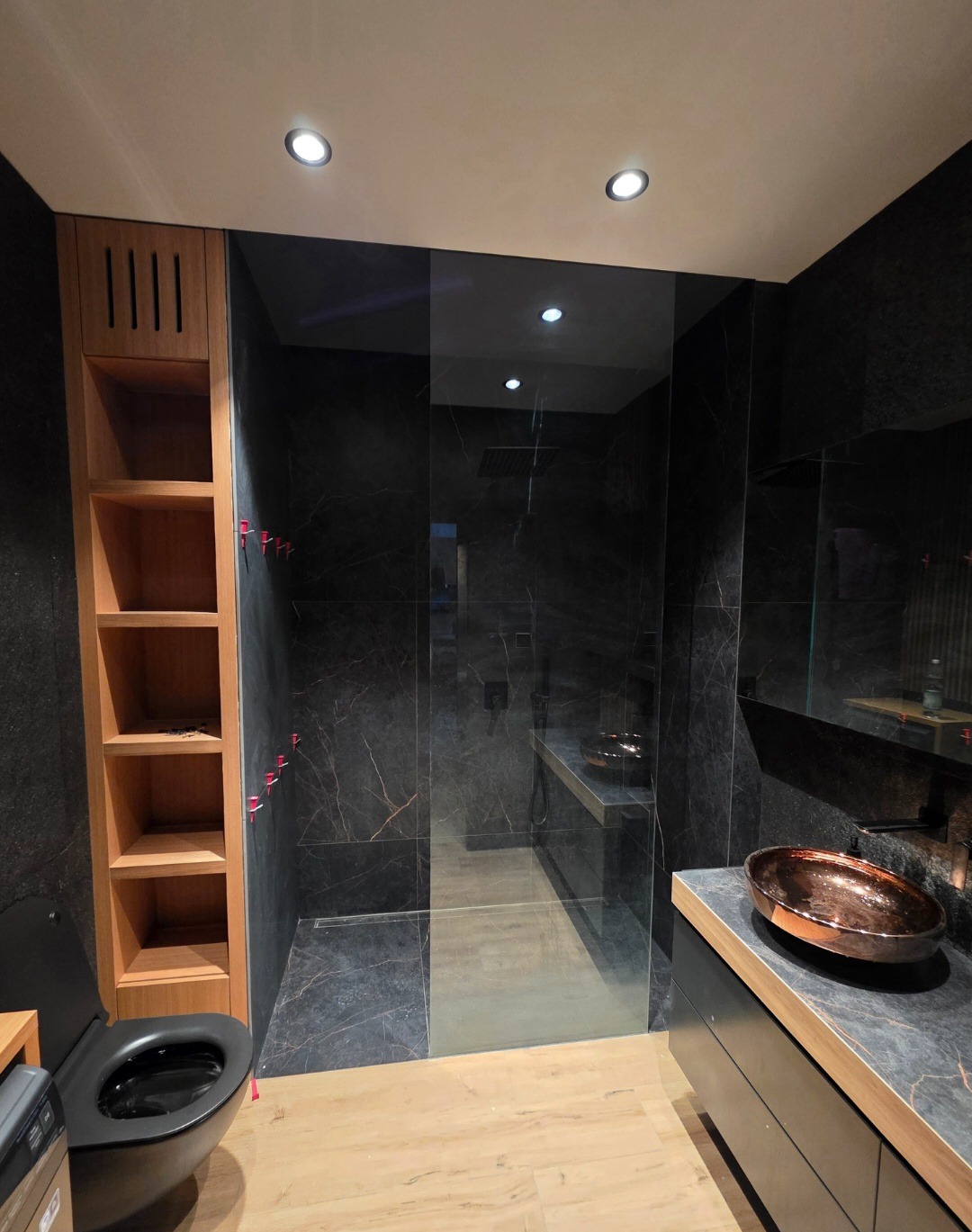 Nowoczesna łazienka z czarnymi marmurowymi ścianami, szklaną kabiną prysznicową i drewnianymi elementami. Uwagę przykuwa miedziana umywalka.