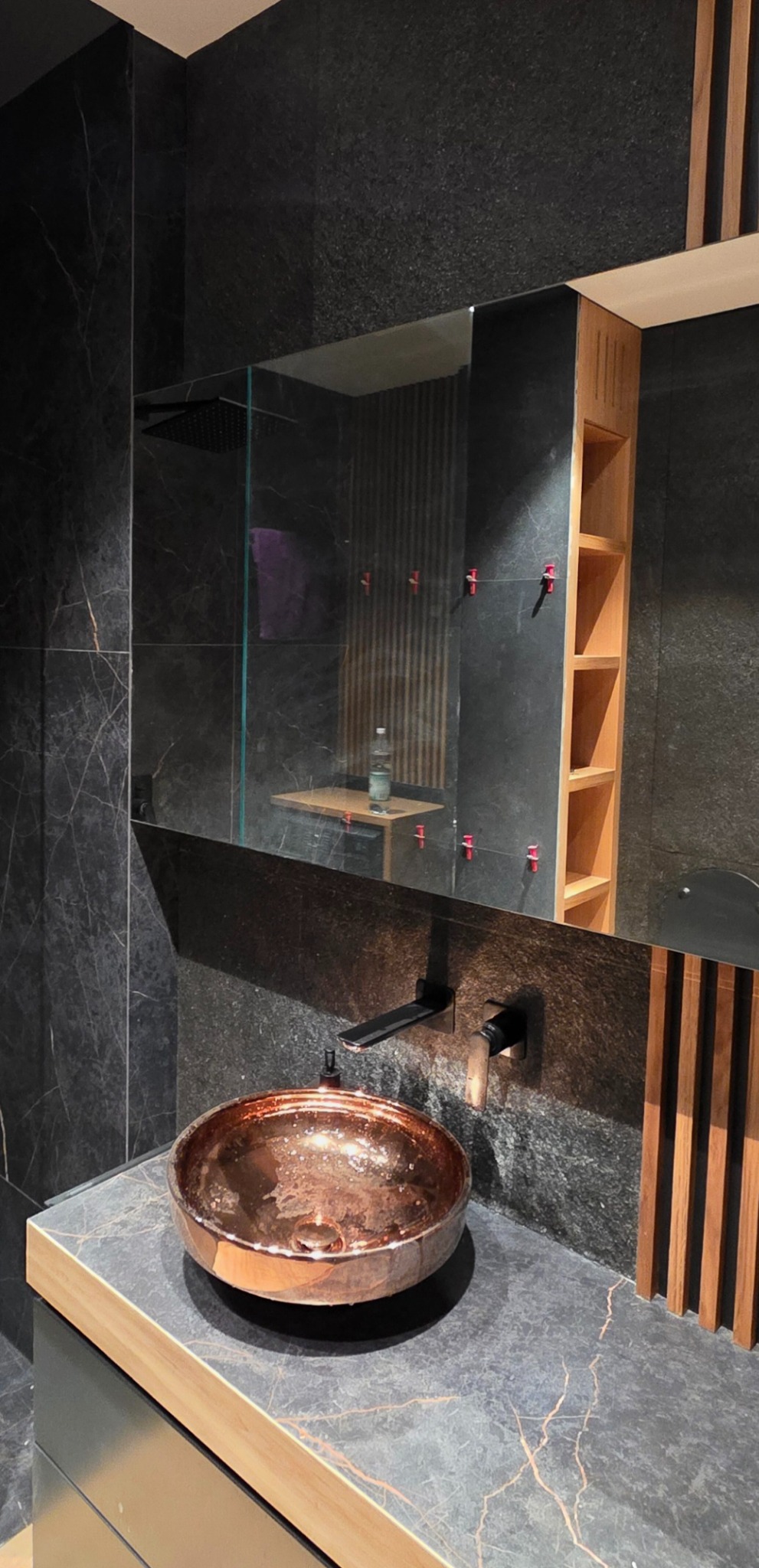 Elegancka łazienka z umywalką w kolorze miedzi, czarnymi płytkami imitującymi marmur i drewnianymi akcentami. W lustrze odbija się prysznic typu deszczownica.
