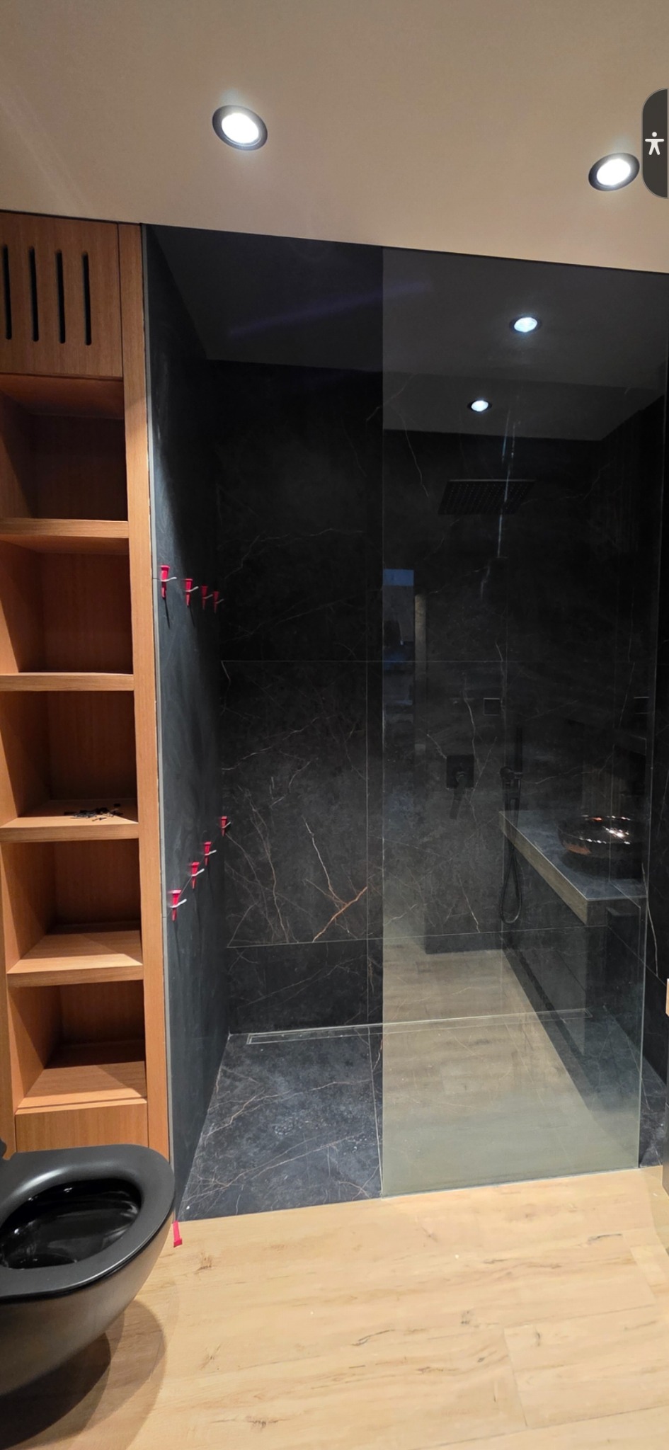 Nowoczesna łazienka z czarnymi marmurowymi płytkami, szklaną kabiną prysznicową i drewnianymi półkami. Widoczne czerwone krzyżyki do poziomowania płytek.