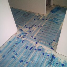 K.m.r - Rozłożona mata grzewcza firmy Mapei z niebieskimi przewodami i taśmą mocującą, przygotowana do wylewki w trakcie prac wykończeniowych w pomieszczeniu.