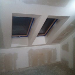 K.m.r - Pokój na poddaszu w trakcie prac wykończeniowych: dwa okna dachowe zabezpieczone taśmą malarską, ściany z widocznymi śladami szpachlowania, przygotowane do malowania.