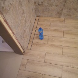 K.m.r - Ukończona podłoga w łazience z płytek imitujących drewno, z widocznym odpływem liniowym i butelką wody mineralnej. Częściowo widoczna ściana wyłożona płytkami ceramicznymi w odcieniach beżu.