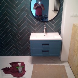 K.m.r - Niedokończona łazienka z zielonymi płytkami ułożonymi w jodełkę, niebieską szafką z umywalką, okrągłym lustrem z odbiciem osoby robiącej zdjęcie, oraz widocznymi materiałami zabezpieczającymi...