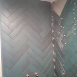 Turkusowe płytki ułożone we wzór jodełki na podłodze w łazience, z widocznymi krzyżykami dystansowymi i dwoma czerwonymi papilotami do włosów.