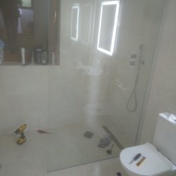 K.m.r - Montaż kabiny prysznicowej w łazience z widocznym odpływem liniowym, narzędziami (wiertarka, śrubokręt) i toaletą w trakcie prac wykończeniowych.