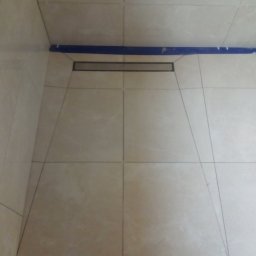 K.m.r - Ułożone płytki podłogowe w kabinie prysznicowej z odpływem liniowym, zabezpieczone niebieską taśmą malarską na styku ze ścianą, widoczne fugi i fragment buta w dolnej części kadru.