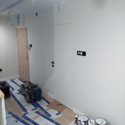 K.m.r - Przygotowania do malowania ścian w pomieszczeniu: zabezpieczona podłoga, narzędzia malarskie, puszki z farbą i białe ściany z zamontowanymi gniazdkami i oświetleniem sufitowym.