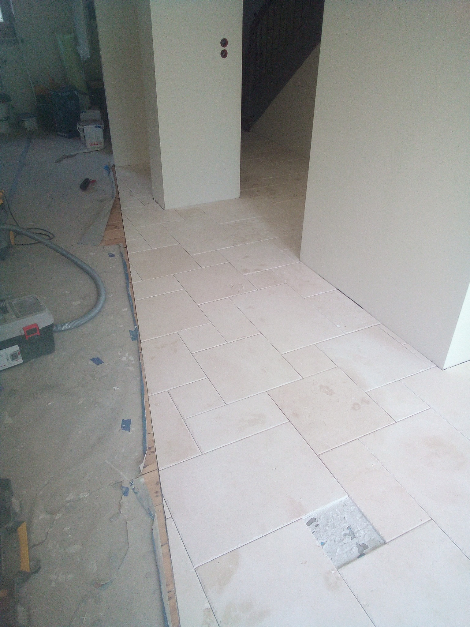 Układanie beżowych płytek podłogowych w korytarzu, widoczny fragment nieukończonej podłogi z brakiem jednej płytki, narzędzia glazurnicze i odkurzacz przemysłowy w tle.