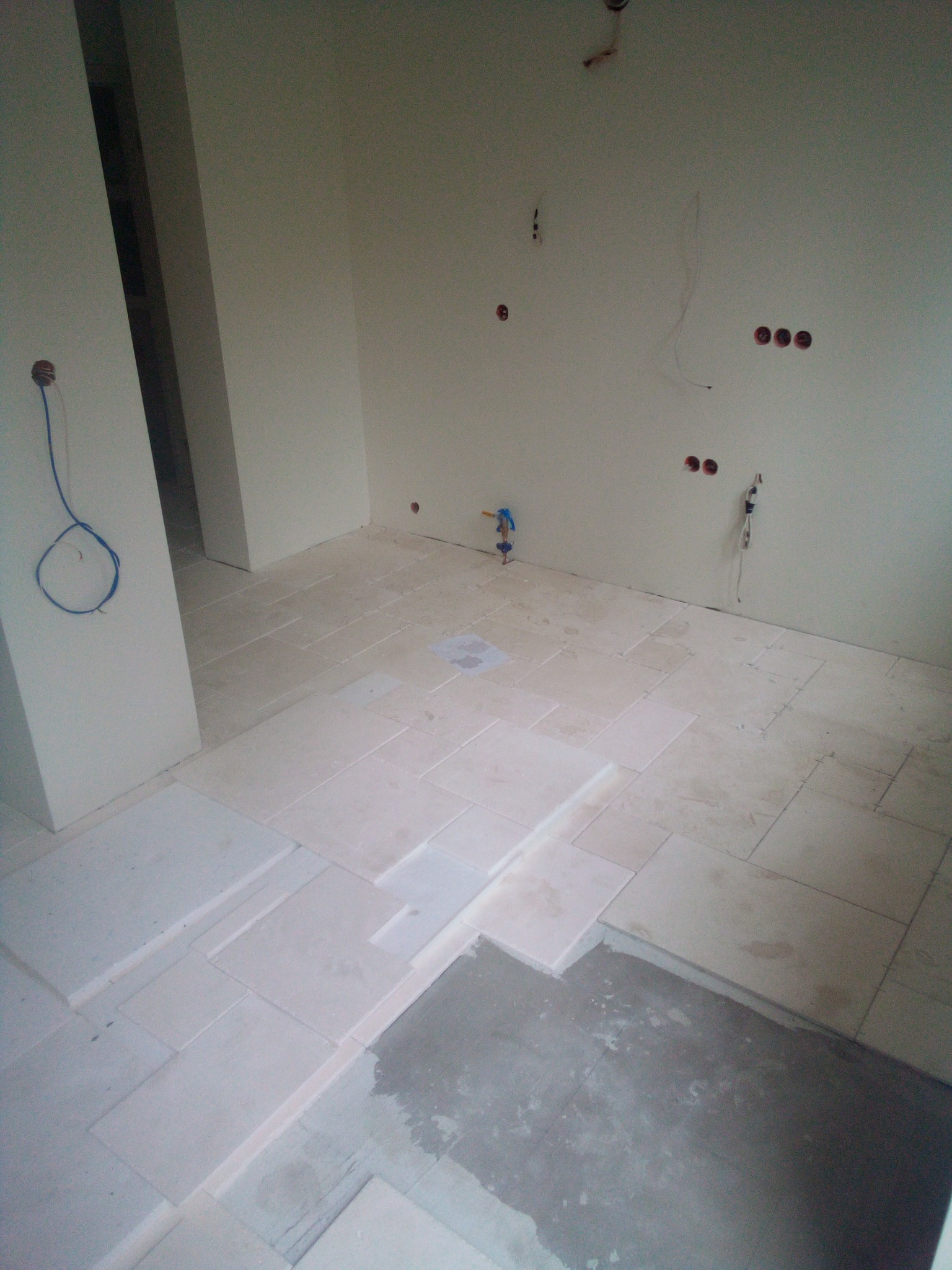 Układanie jasnych, prostokątnych płytek na podłodze w trakcie prac wykończeniowych, widoczne odsłonięte przewody elektryczne i rury doprowadzające wodę.