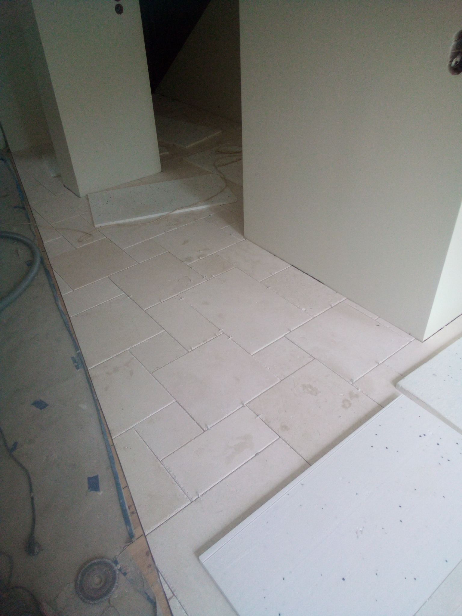 Układanie beżowych płytek podłogowych w korytarzu, widoczne fragmenty niezakończonej pracy i płyty styropianowe.