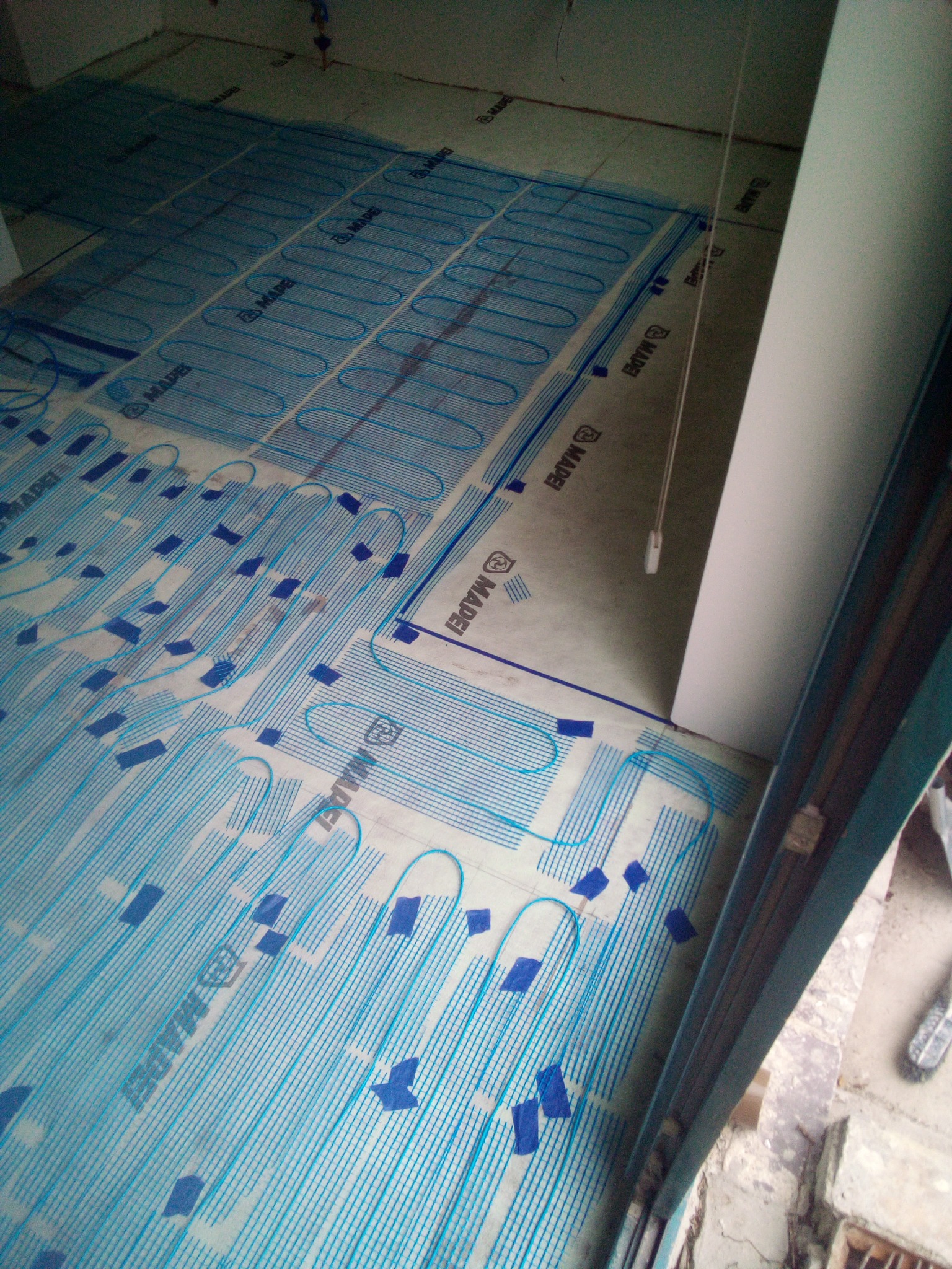 Rozłożona mata grzewcza podłogowa Mapei, przygotowana do wylewki, widok z góry, fragment okna w prawym dolnym rogu.