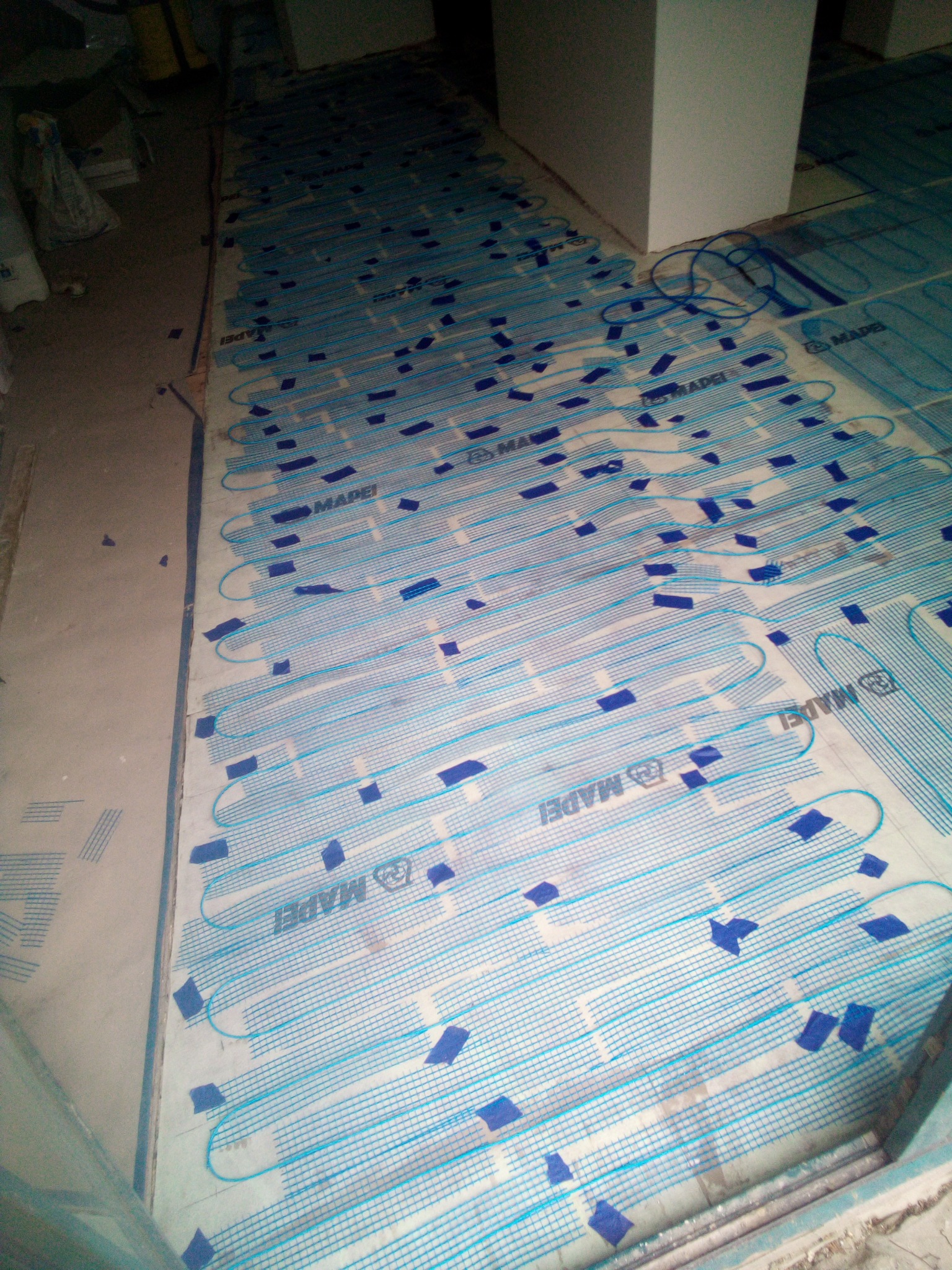 Rozłożona mata grzewcza Mapei z niebieskimi przewodami i niebieską taśmą mocującą na podłodze w trakcie prac przygotowawczych, widoczne kable zasilające.