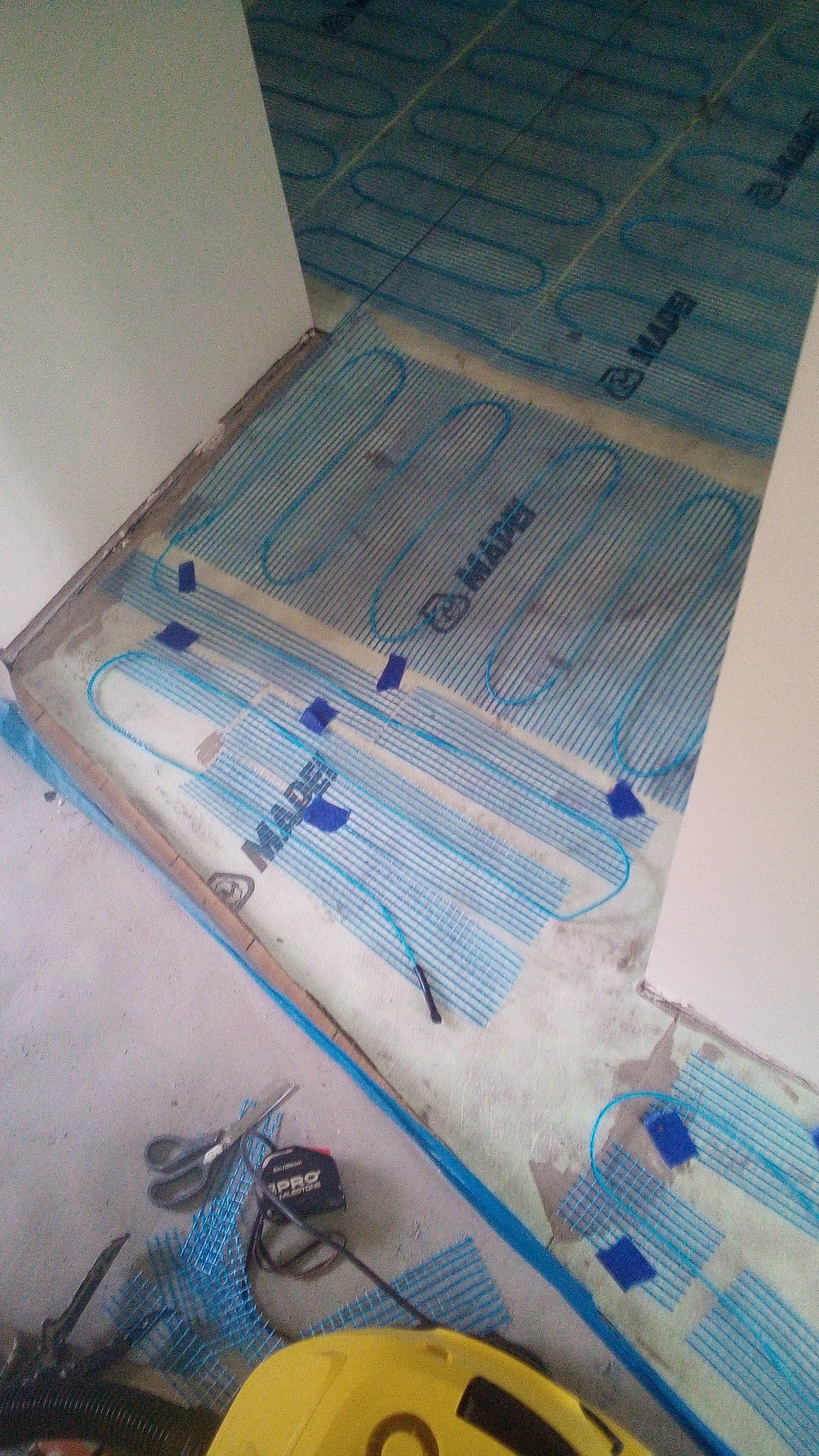 Rozłożona mata grzewcza Mapei na podłodze w trakcie montażu, przytwierdzona niebieską taśmą, widoczne narzędzia: nożyczki, miarka i odkurzacz.