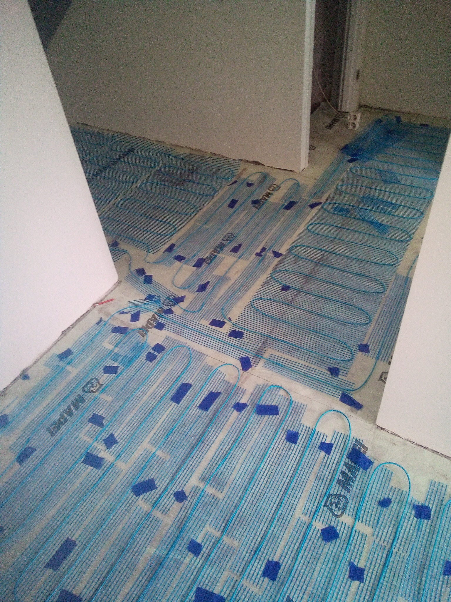 Rozłożona mata grzewcza firmy Mapei z niebieskimi przewodami i taśmą mocującą, przygotowana do wylewki w trakcie prac wykończeniowych w pomieszczeniu.