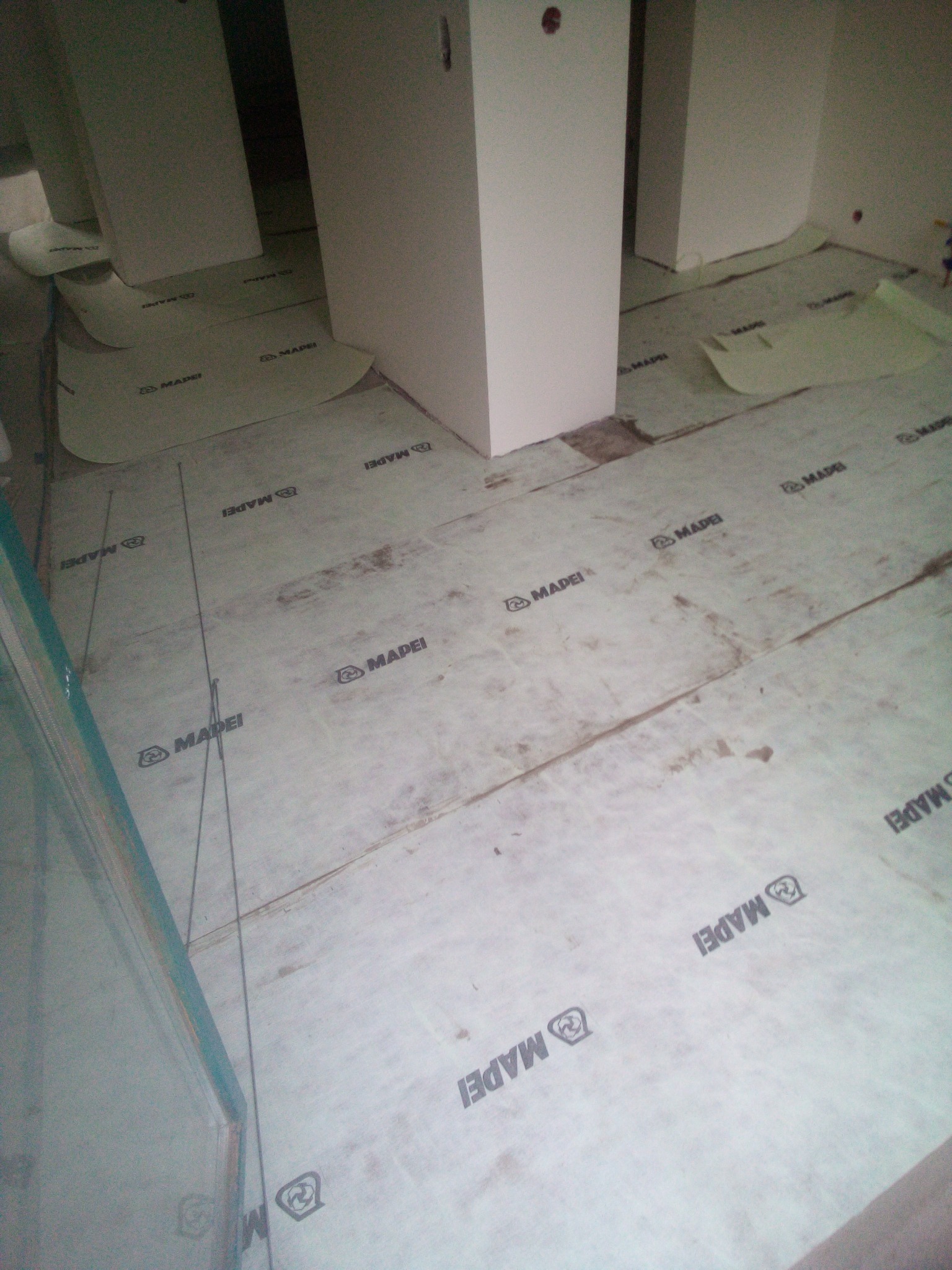 Przygotowanie podłoża pod płytki w łazience, widoczne płyty podkładowe z logo Mapei, pionowe linie traserskie i fragment zabudowy meblowej.