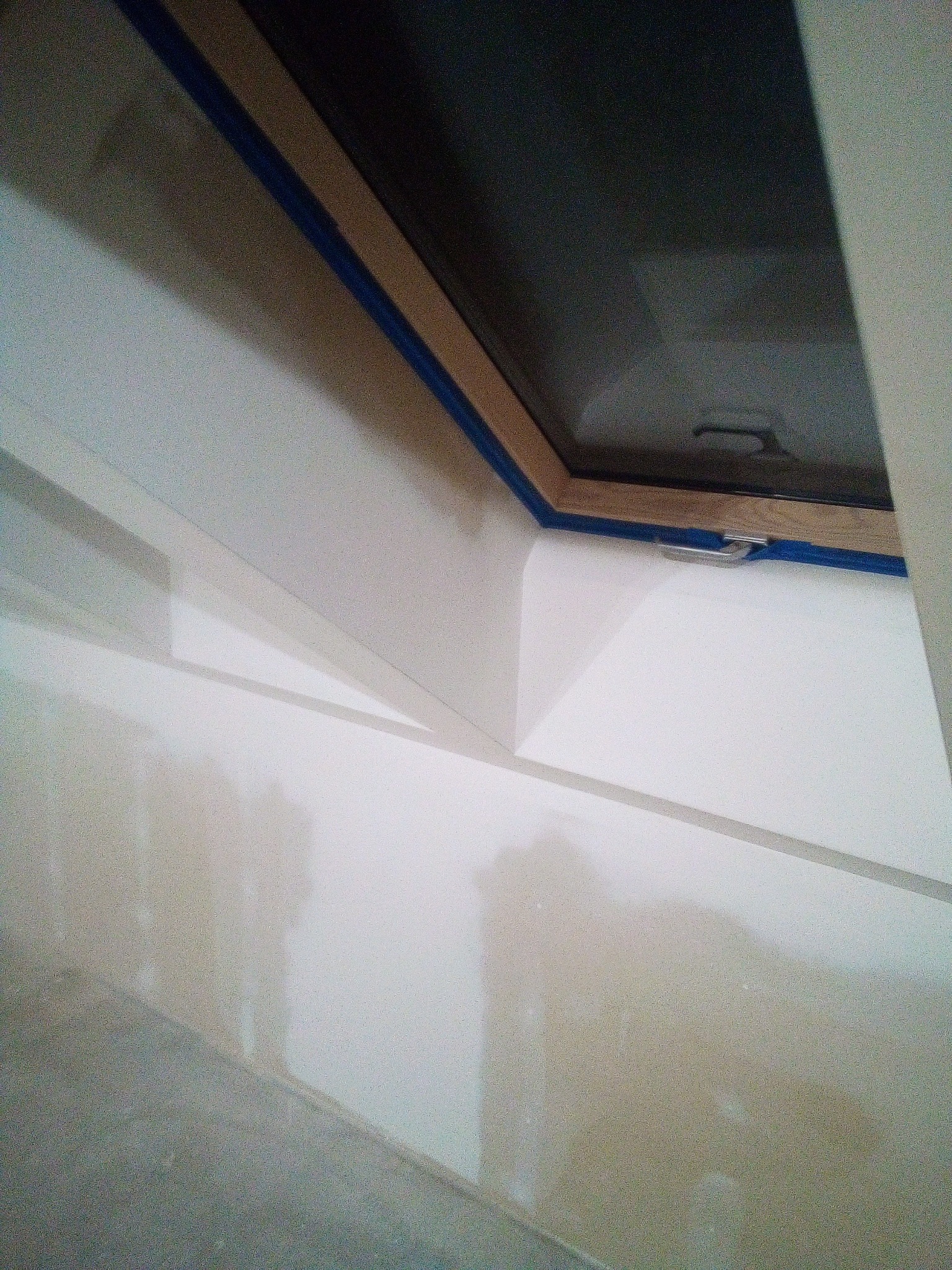 Okno dachowe z drewnianą ramą zabezpieczoną niebieską taśmą malarską, osadzone w białej ścianie, widoczne ślady szpachlowania.